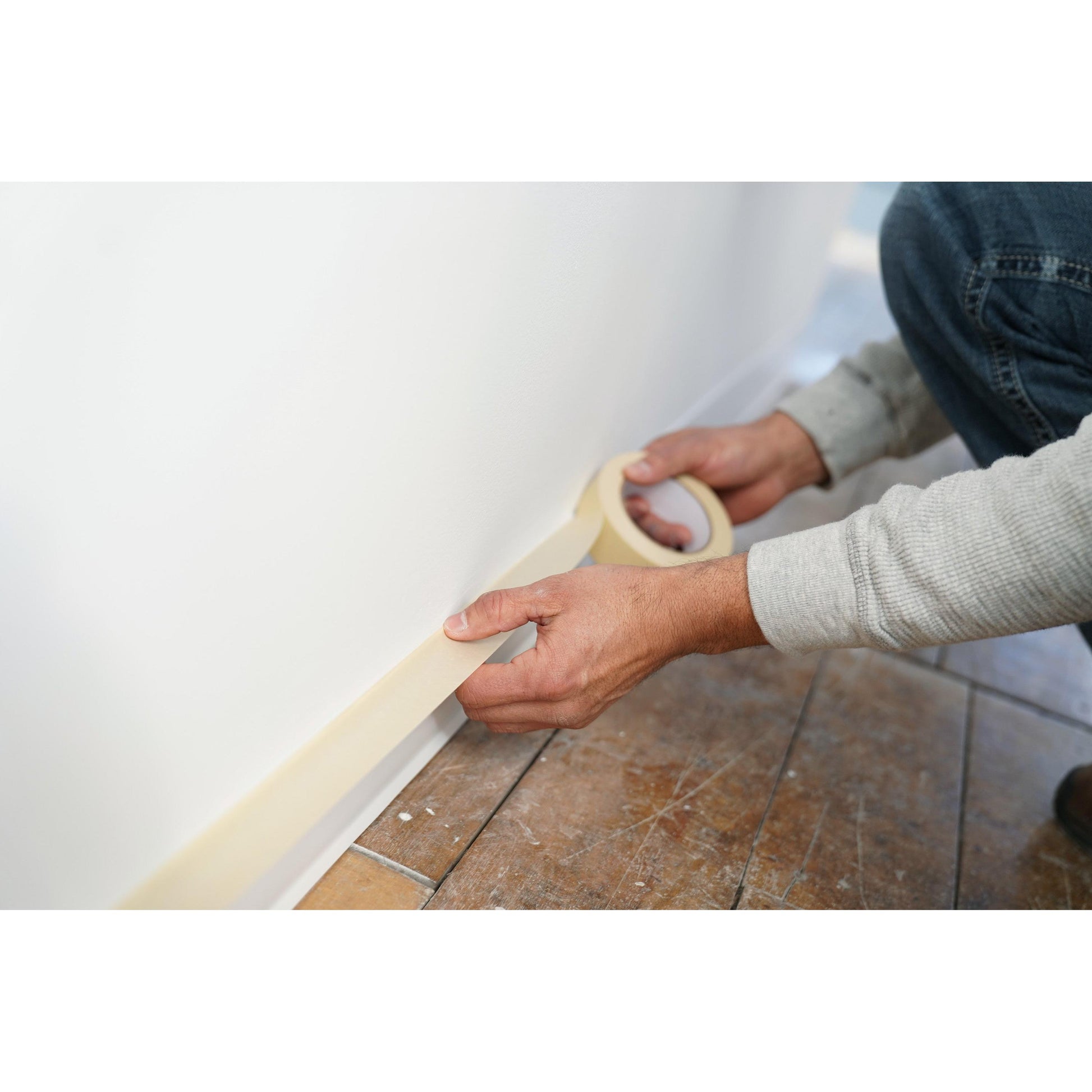 Eine Person in einem langärmeligen Hemd klebt Scotch® Classic Abdeckband (24 mm x 50 m, 1 Rolle) von 3M Deutschland GmbH entlang der Unterkante einer weißen Wand in der Nähe eines holzgemusterten Fußbodens, wahrscheinlich als Vorbereitung für Malerarbeiten.
