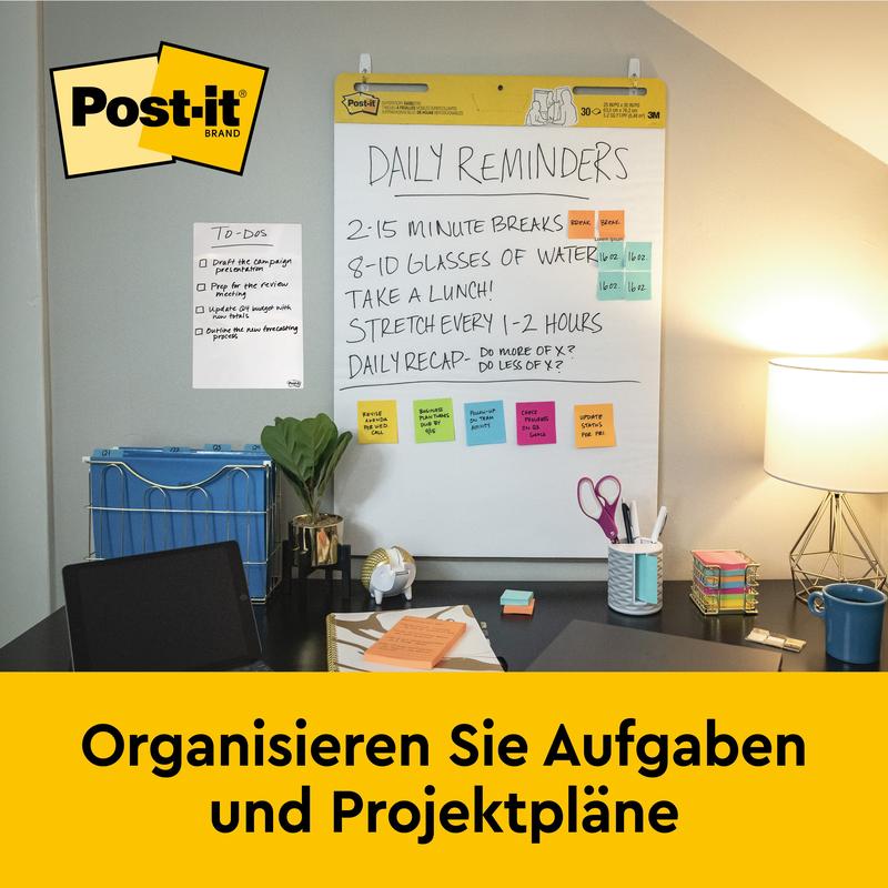Et skrivebord med kontorartikler, lampe og stol står foran en vægmonteret 3M Post-it® Super Sticky selvklæbende mødeplanchart (63,5x76,2 cm, 30 ark), der viser daglige påmindelser og noter. På et gult banner nedenunder står der "Organiser opgaver og projektplaner.