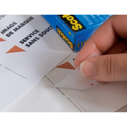 Eine Hand zieht einen dreieckigen Aufkleber von einem Blatt mit der Aufschrift "IMAGE DE MARQUE SERVICE SANS SOUCI" ab, während im Hintergrund eine Schachtel 3M Deutschland GmbH Scotch® unsichtbares Klebeband, ablösbar, 19 mm x 33 m, 1 Rolle | 1 Stück steht.