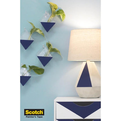 En blå-hvid, geometrisk kommode og en lampe står foran en blå væg med trekantede plantekasser, alt beskyttet med Scotch® malertape til sarte overflader (36 mm x 41 m, 1 rulle) fra 3M Deutschland GmbH. Logoet vises nederst til venstre.
