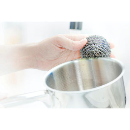 En hånd bruger en Scotch-Brite® rustfrit stål spiral fra 3M Deutschland GmbH, 3 stykker pr. pakke, til at fjerne genstridige pletter ved rengøring af en rustfri stålgryde.