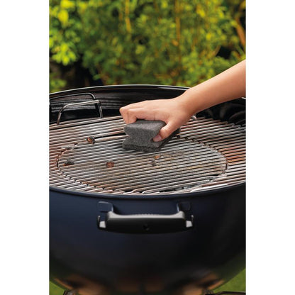 En hånd rengør metalgrillen på en rund kulgrill med en Scotch-Brite® Grillgrebsvamp (1 styk pr. pakke) fra 3M Deutschland GmbH. Grønne blade er slørede i baggrunden.