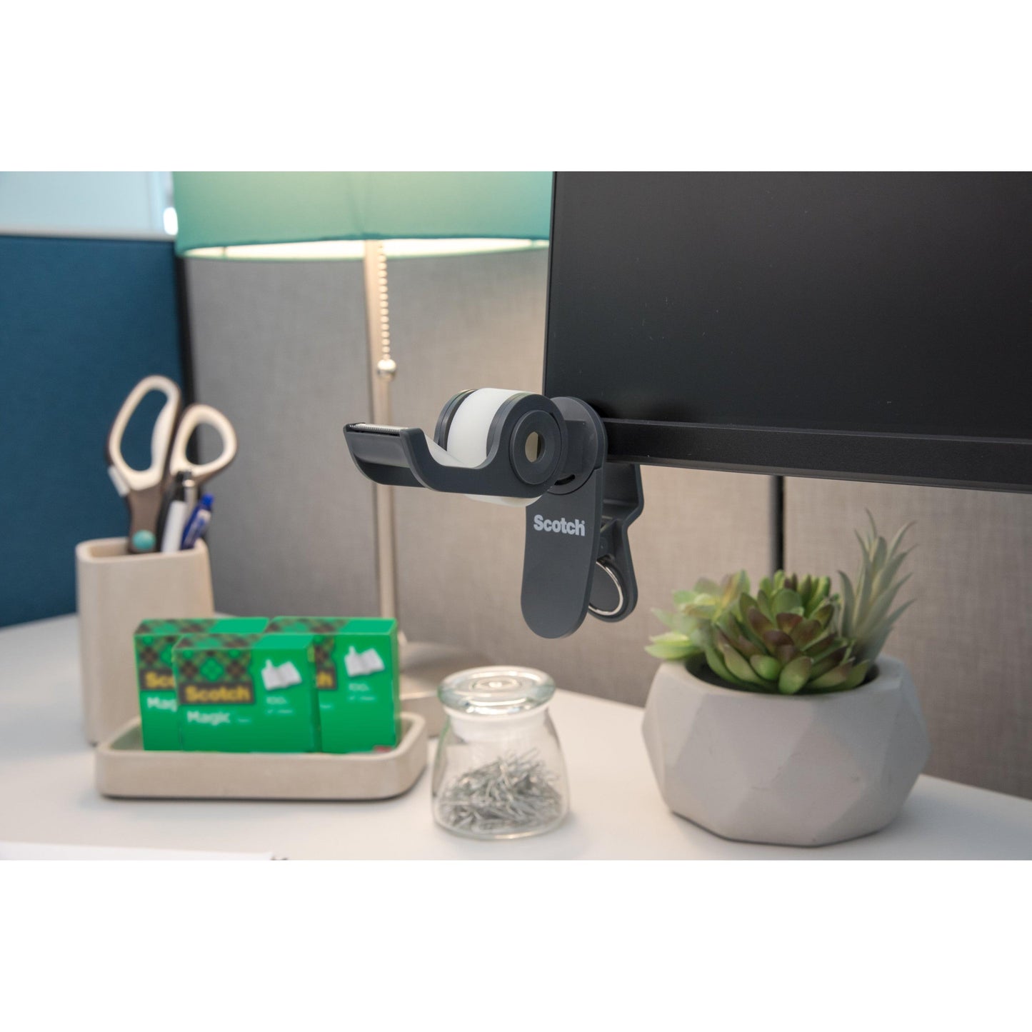 Auf dem Schreibtisch: ein 3M Scotch™ Clip & Twist-Klebebandabroller mit Scotch® Magic™ Invisible Tape (19 mm × 8,89 m), eine Lampe, eine Schere in einem Halter, zwei zusätzliche Rollen Scotch®-Klebeband, ein Glas mit Stecknadeln und eine kleine Sukkulentenpflanze im Topf.
