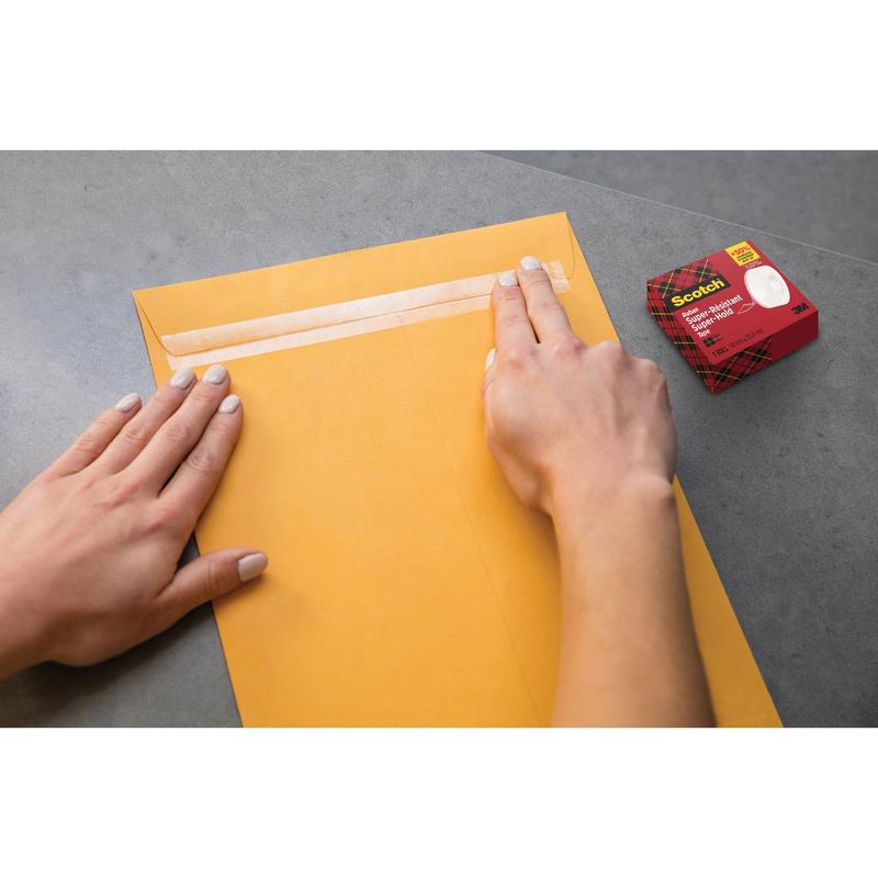 En person forsegler en stor brun konvolut med Scotch® Super-Hold Secure Tape, 1 rulle, 19 mm x 25,4 m fra 3M Deutschland GmbH på en grå overflade. Tapens kasse er i nærheden og sikrer en stærk og gennemsigtig forsegling til sikker postforsendelse.