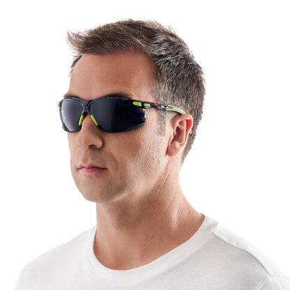 En mand med kort brunt hår bærer en hvid T-shirt med rund halsudskæring og en 3M™ Solus™ beskyttelsesbrille 1000 solbrille (grøn/sort, gråt glas, Scotchgard™ antidug-belægning) fra 3M Deutschland GmbH foran en enkel hvid baggrund.