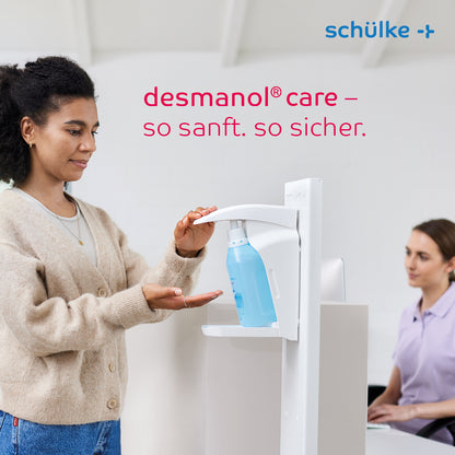 En kvinde bruger en Schülke desmanol® care hånddesinfektionsmiddel-dispenser ved en skranke, mens en anden kvinde sidder i nærheden og arbejder på en computer. Teksten "desmanol® care – så mild. så sikker." og mærket "Schülke & Mayr GmbH" præsenteres, hvor hudplejeformlen for mild og effektiv anvendelse af hånddesinfektionsmiddel er i fokus.